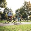 Two older people with white hair ride bicycles on a path through a park.