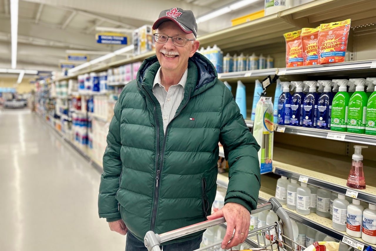 A tall man with a moustache and a ball cap smiles at the camera in a grocery store with his hand on his cart handle. 