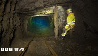 A man is a yellow boilersuit stands in an old mine working, the rocky tunnel ends in a green blue pool of water