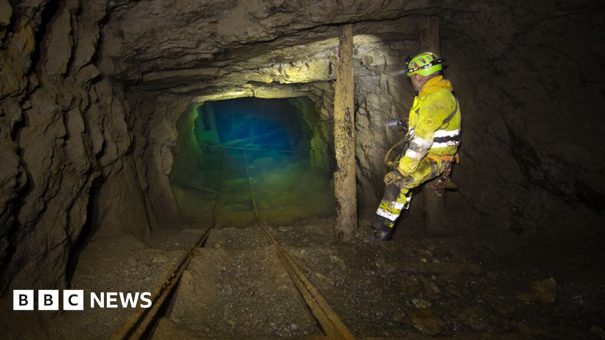 A man is a yellow boilersuit stands in an old mine working, the rocky tunnel ends in a green blue pool of water