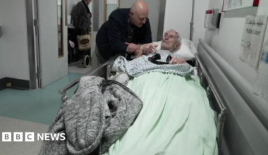A man leans over an elderly patient lying on a trolley in a hospital corridor, with other patients and equipment visible in the background.