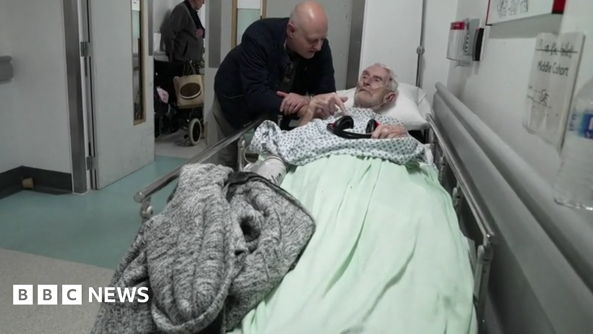A man leans over an elderly patient lying on a trolley in a hospital corridor, with other patients and equipment visible in the background.