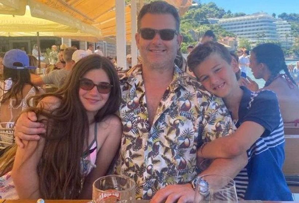 James Molloy with children Jasmine and Edward, all smiling and posing for a photograph, are seated at an outdoor dining area, with a blurred background of other diners and a distant landscape.