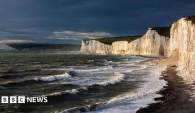 Waves crash to the shore beside some large white cliffs. The sky looks dark and stormy.