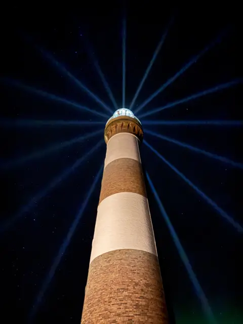 Cameron Reid A tall brick chimney stands against a dark night sky with blue light beams fanning out behind it.