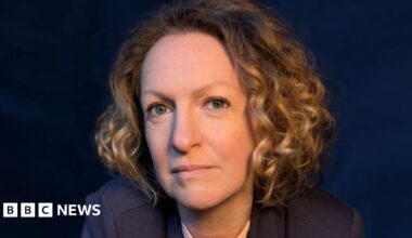 A professional headshot of Victoria Woodall with chin length golden curly hair wearing a navy suit against a navy backdrop
