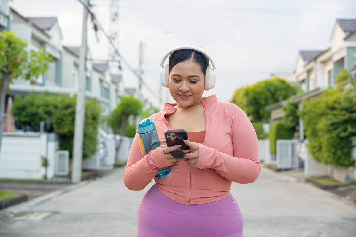 Beautiful-plus-size-woman-in-sportswear-holding-water-bottle-using-smartphone-and-headphones-after-workout-in.jpg