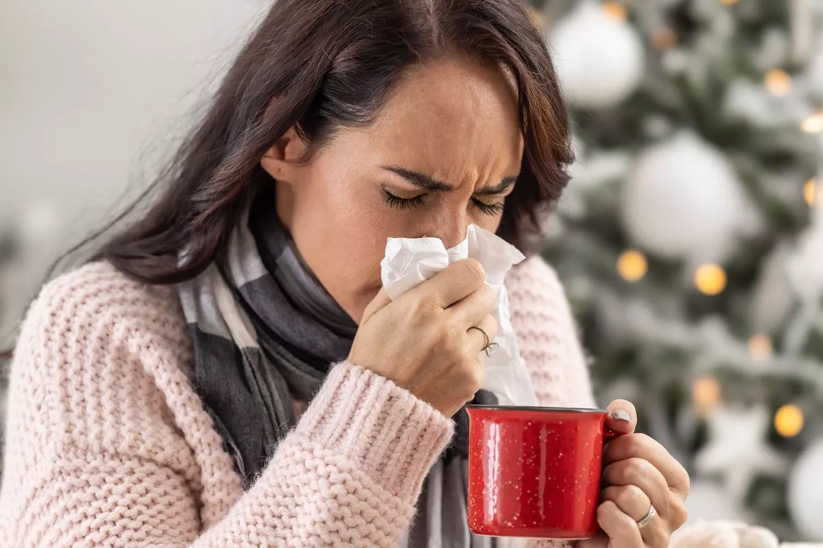 Woman experiencing cold symptoms and sneezing dressed with a blanket drinking hot tea.