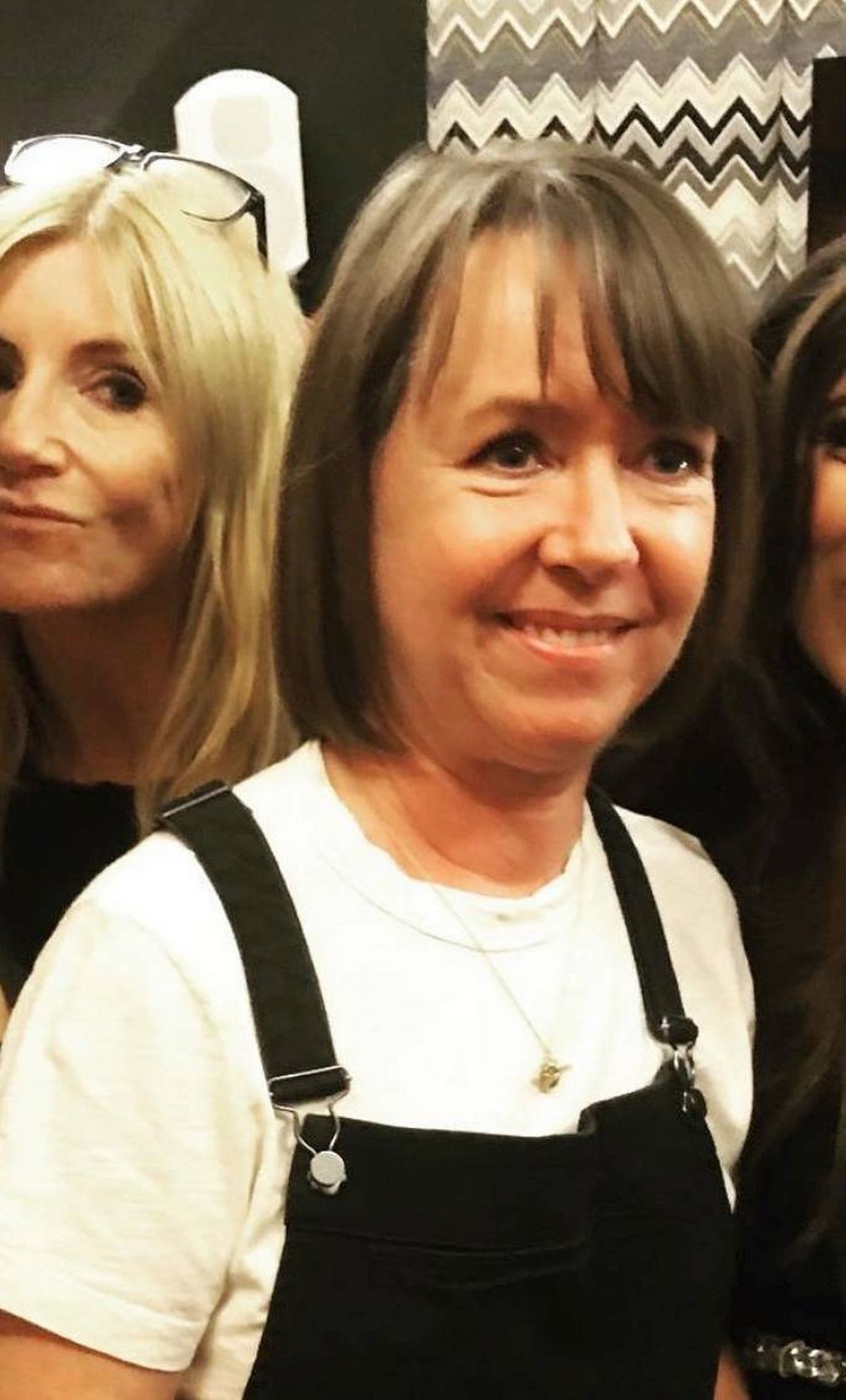 An image depicts three individuals standing close together, possibly at an event. The woman in the center has short, dark hair and is smiling broadly. To her left, another woman with long, light blonde hair is also smiling. In the background, a third woman with partially visible dark hair is present.