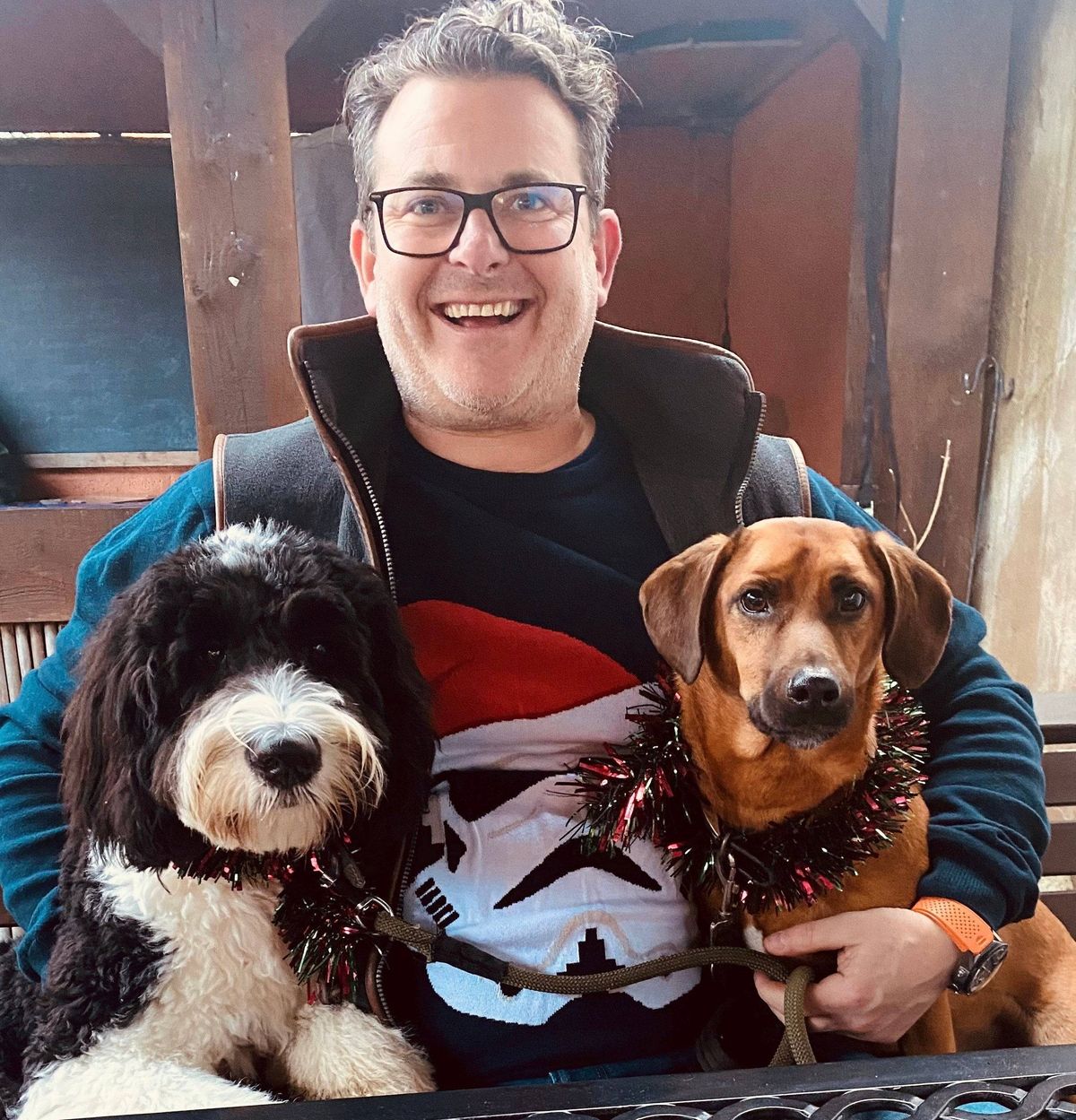 An individual wearing a blue jacket and glasses stands with two dogs, one brown and one black and white. The person is smiling and holding the leashes of the dogs. The background appears to be a wooden structure.