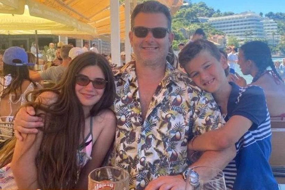 An adult male and two children, all smiling and posing for a photograph, are seated at an outdoor dining area, with a blurred background of other diners and a distant landscape.