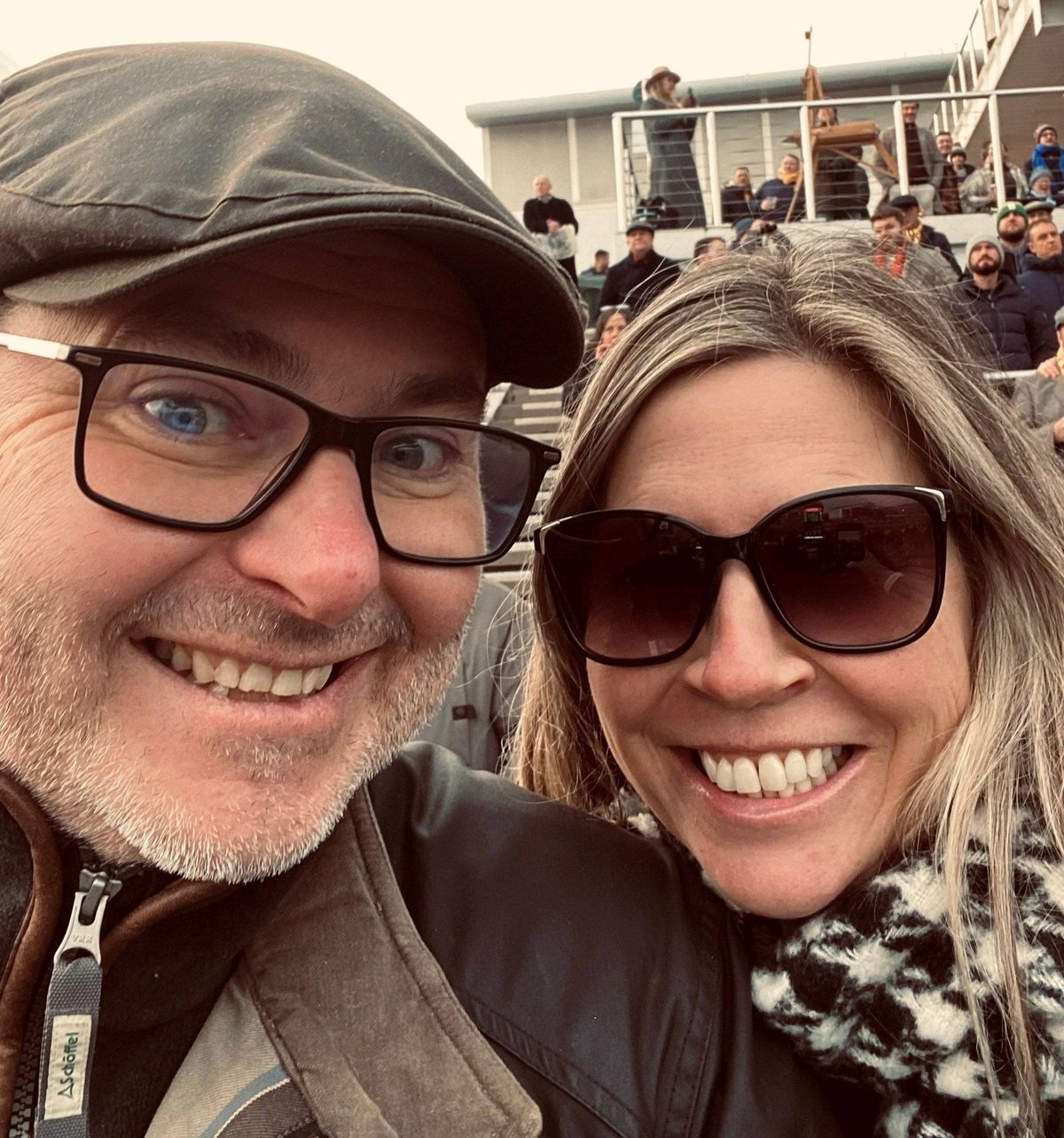 An adult male wearing a fedora and glasses, and an adult female wearing sunglasses and a scarf are smiling and posing for a photo in an outdoor setting with a crowd of people in the background.