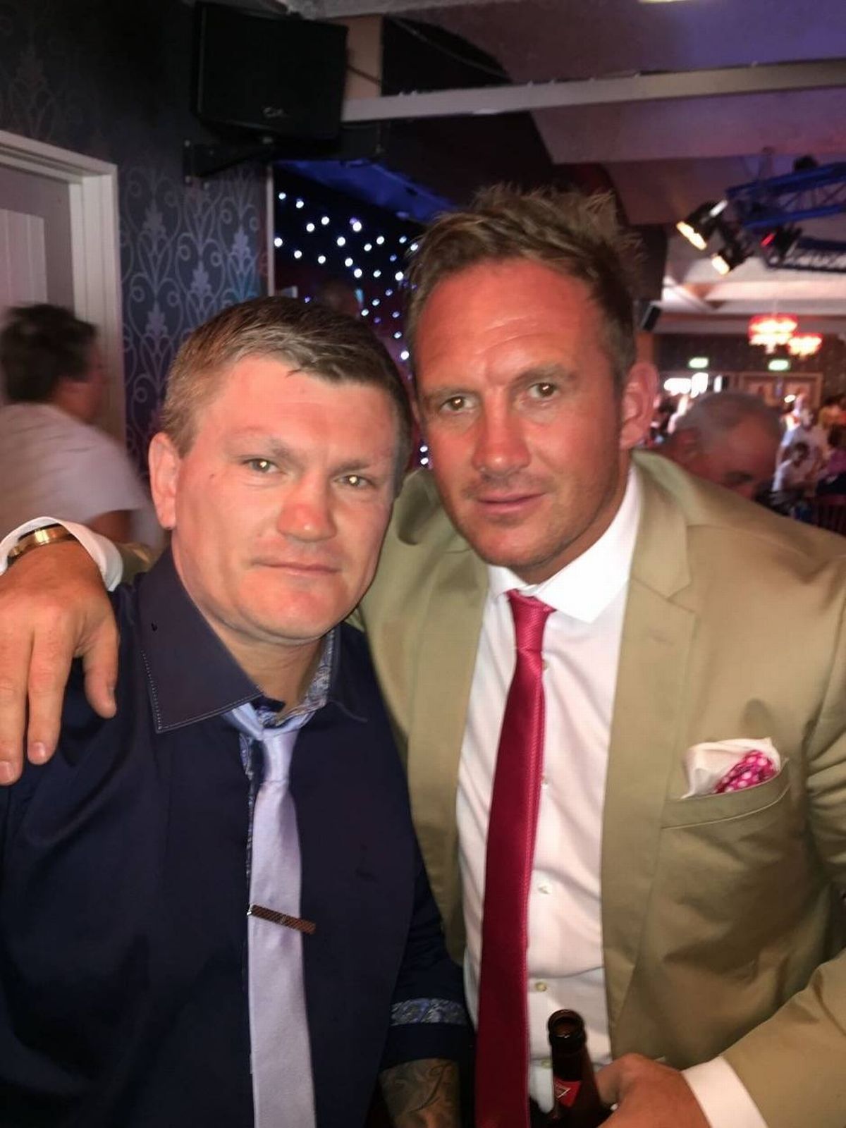 Two men dressed formally are standing next to each other, possibly at a social event. One man is wearing a beige suit and a red tie, while the other man is dressed in a dark suit with a blue tie. They appear to be posing for a photograph. In the background, several people can be seen attending to what seems to be an event or gathering.