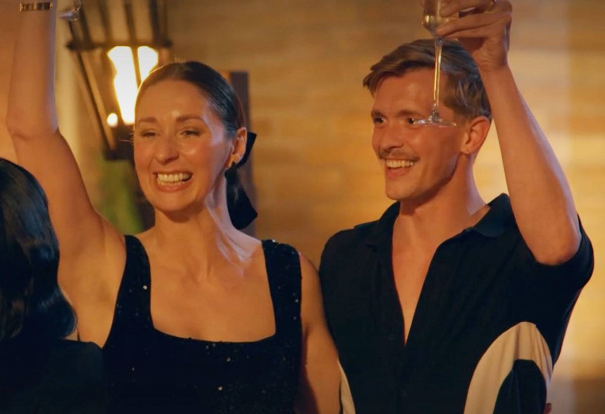 An image depicts two individuals, both dressed in black attire, standing close to each other with their arms raised and expressions of joy on their faces, suggesting a moment of celebration or performance. The setting appears to be indoors, with ambient lighting enhancing the festive atmosphere.