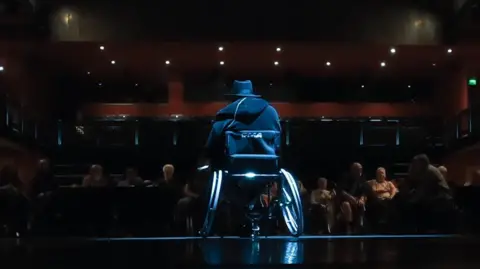 Supplied The back of a man in a wheelchair on a stage in front of an audience