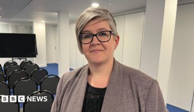 Deputy Haley Camp - A woman with grey hair, wearing black rimmed glasses. She has a grey checked jacket on over a black blouse. She is standing in front of rows of chairs, organised for a presentation.