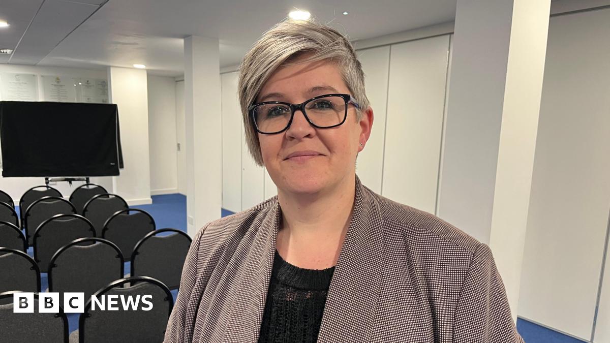 Deputy Haley Camp - A woman with grey hair, wearing black rimmed glasses. She has a grey checked jacket on over a black blouse. She is standing in front of rows of chairs, organised for a presentation.