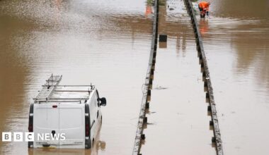 A white van is stranded in floodwater on the A189 Spine Road near Blyth, Northumberland. The water comes up to the the top of the vehicle's number plate. A worker in an orange high-vis uniform is crouched down further up the road.
