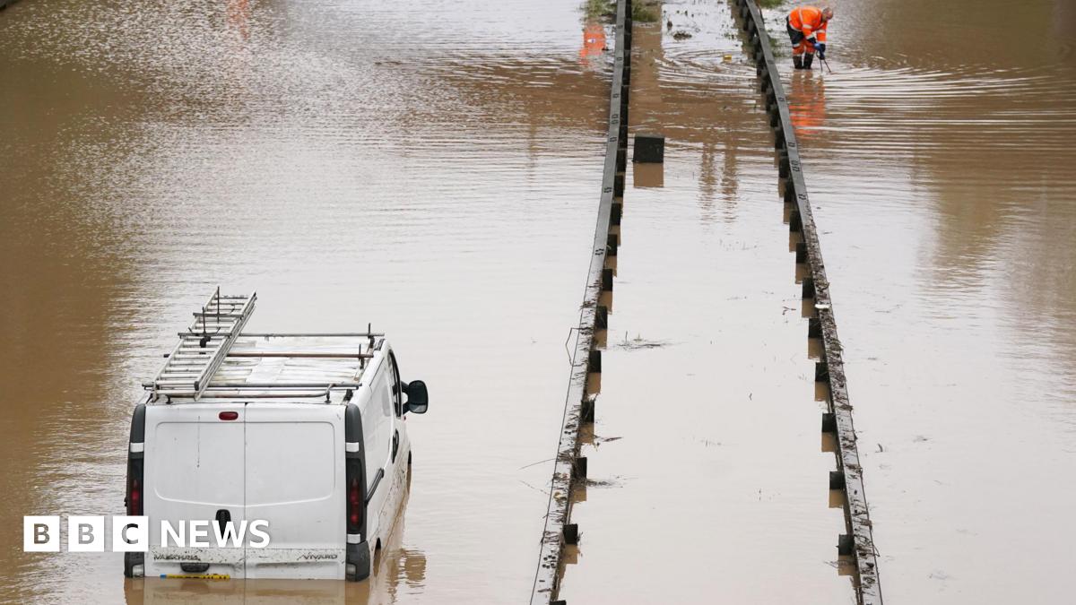 A white van is stranded in floodwater on the A189 Spine Road near Blyth, Northumberland. The water comes up to the the top of the vehicle's number plate. A worker in an orange high-vis uniform is crouched down further up the road.