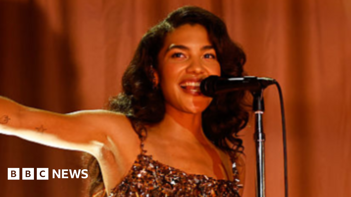 Dean with long, wavy dark hair wearing a sparkly flapper dress, singing into a microphone with one arm stretched out to the side, pointing. Behind her is a gold curtain.