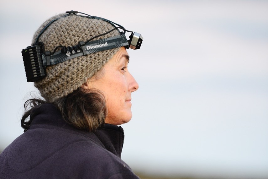 A woman in a beanie and a head torch looks into the distance.