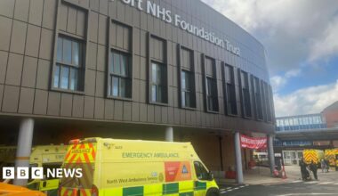 An ambulance at the entrance to Stepping Hill hospital in Stockport, with the main hospital building above a roadway and entrance signs and buildings.