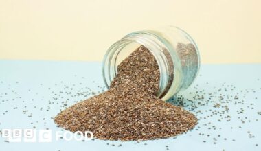 A glass jar on its side, which is full of seeds including chia seeds which are spilling out on a blue and pale yellow background