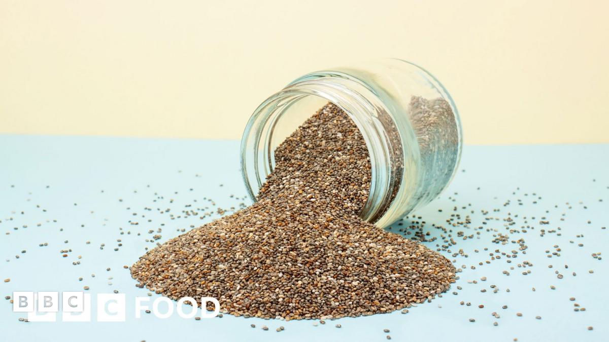 A glass jar on its side, which is full of seeds including chia seeds which are spilling out on a blue and pale yellow background