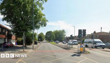 Daytime image of the A34 Kingsway in Manchester. There is no traffic on the left hand side of the road but several vehicles on the right. A woman holding a small child with a pram nearby stands on the pavement to the left of a yellow junction.