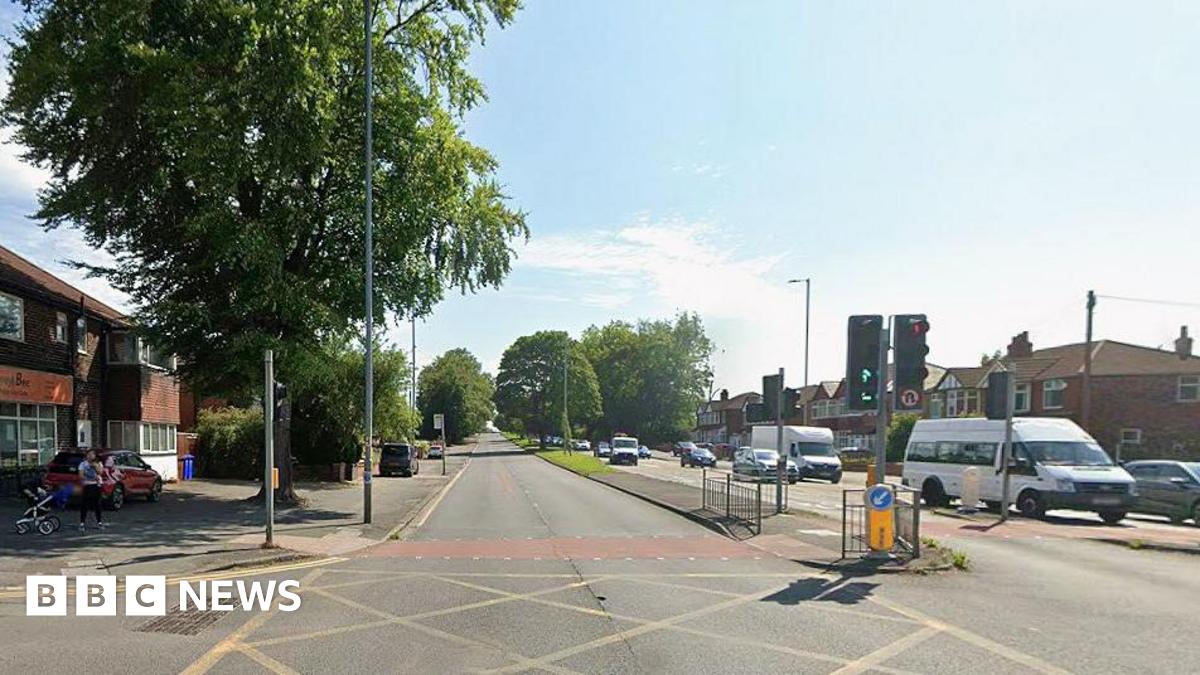 Daytime image of the A34 Kingsway in Manchester. There is no traffic on the left hand side of the road but several vehicles on the right. A woman holding a small child with a pram nearby stands on the pavement to the left of a yellow junction.