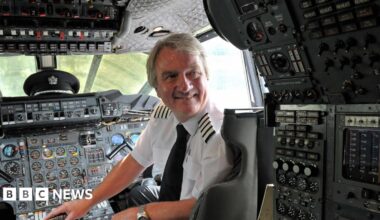 John Tye standing in front of Concorde plane Alpha Foxtrot on the runway. He is dressed in his black pilot uniform and a black cap. He has his hands crossed in front of him. Behind his, others can be seen fixing up the plane, with a bright blue sky above them.