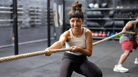 Getty Images A young woman wearing black leggings and a sleeveless top, with dark hair tied up, pulls hard on a tug-of-war rope in a gym with a grey floor. Shelves of weights and are seen in the background, as well as a man (blurred) holding the same rope, wearing red shorts.