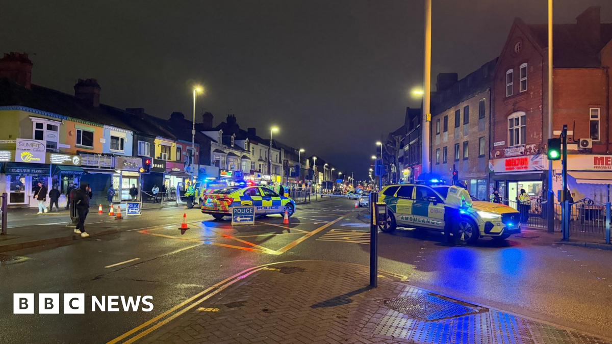 A high street lined with several shops is cordoned off by police. A police car and an ambulance car, both with flashing lights, are in the centre of the road.
