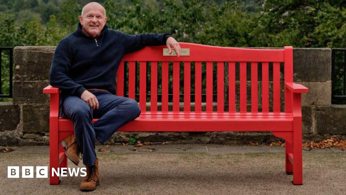 Dean Dyble sits on a bright red wooden red bench. He is an older man, with a bald head and a blue jumper, blue jeans and brown boots. In the background, trees and a stone wall can be seen.