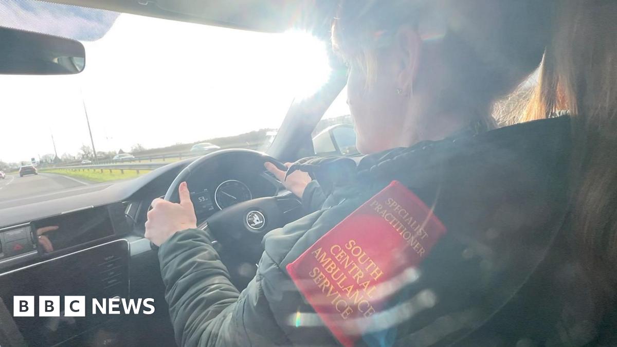 Chrissy Ames is driving her ambulance in the fast lane of a dual carriageway. The sun is shining through the windscreen. Chrissy has both hands on the wheel and her hair tied back in a ponytail. There is gold embroidery on the red epaulette on her shoulder which says specialist practitioner South Central Ambulance Service.