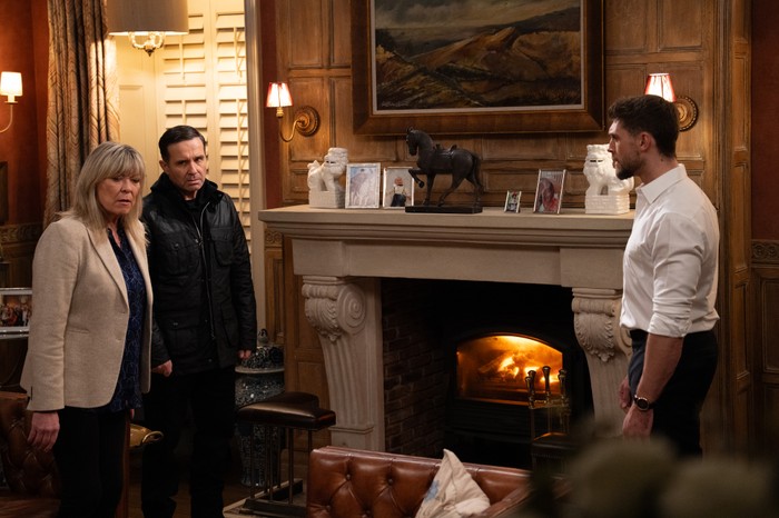 A stunned Joe stands opposite Kim and Graham beside the Home Farm fireplace in a scene from Emmerdale