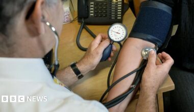 A doctor takes the blood pressure of a patient and listens to their pulse with a stethoscope. A black telephone stands on the desk in the background.