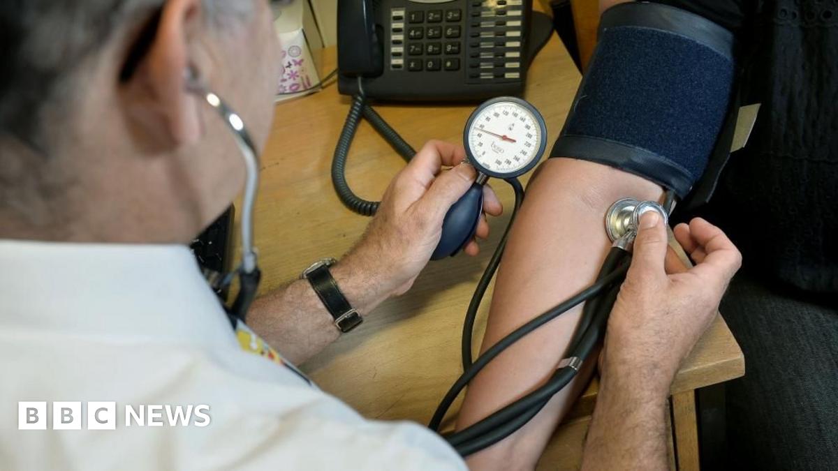 A doctor takes the blood pressure of a patient and listens to their pulse with a stethoscope. A black telephone stands on the desk in the background.