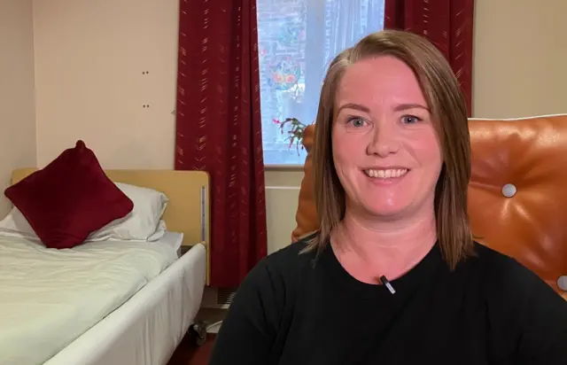 Woman in a care home room.She has brown hair and is wearing a black top. There's a single bed behind her and it has a red cushion on it. There are red curtains around the window.