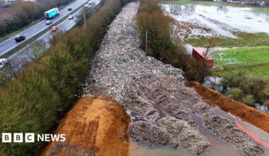 An aerial shot of the dumped waste, stretching out in a road-like line into the distance and surrounded on both sides by trees next to a motorway.