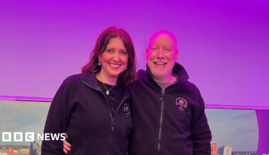 A woman with dark hair hugging a bald man man in a radio studio with a purple backdrop