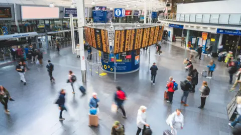 Getty Images An overhead view of Manchester Piccadilly station and a central departures board lit up with train destinations and times. Around thirty people are crossing the concourse, blurred in the photo due to movement.
