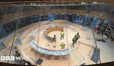 The image shows construction workers at work in the Senedd debating chamber. The politicians' desks are covered in polythene.