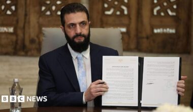 Syrian President Ahmed Sharaa in a navy suit, pale blue tie and white shirt holding up a physical copy of the agreement