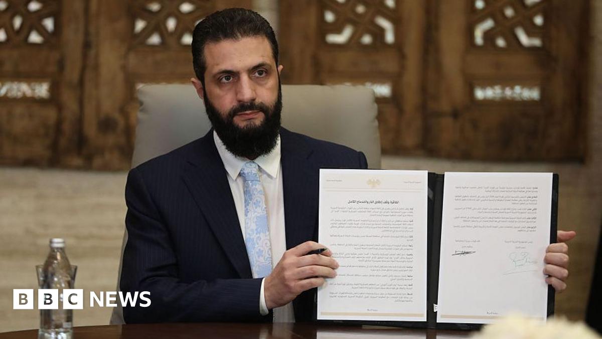 Syrian President Ahmed Sharaa in a navy suit, pale blue tie and white shirt holding up a physical copy of the agreement