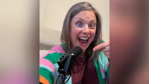 Charlie Flounders A woman with grey and brown hair is smiling and pointing art a black podcast microphone. She is wearing an orange, pink, green and blue cardigan, and has one purple eye