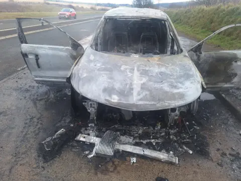 Anne Marie Thomas A front-on view of the the white car completely damaged by the fire. All the windows have gone and there is debris around the vehicle. 