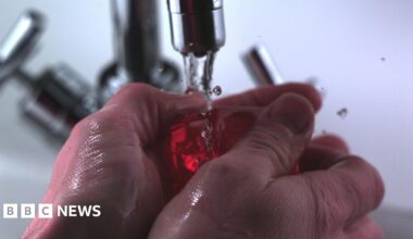 A pair of hands are underneath a tap of running water and are holding a bar of red soap. They are about to lather up the soap.
