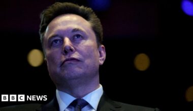 A close-up shot of Elon Musk in a dark room. He stands with a neutral expression against a black background. He has short dark brown hair, and wears a black jacket, white shir,t and dark tie.