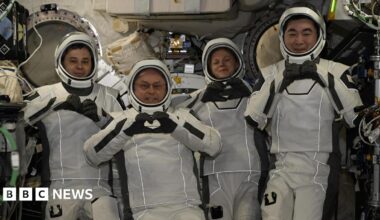 Four astronauts wearing white spacesuits and helmets pose for cameras smiling and making heart shapes with their hands and fingers. They are surrounded by wires and other equipment inside the International Space Station.